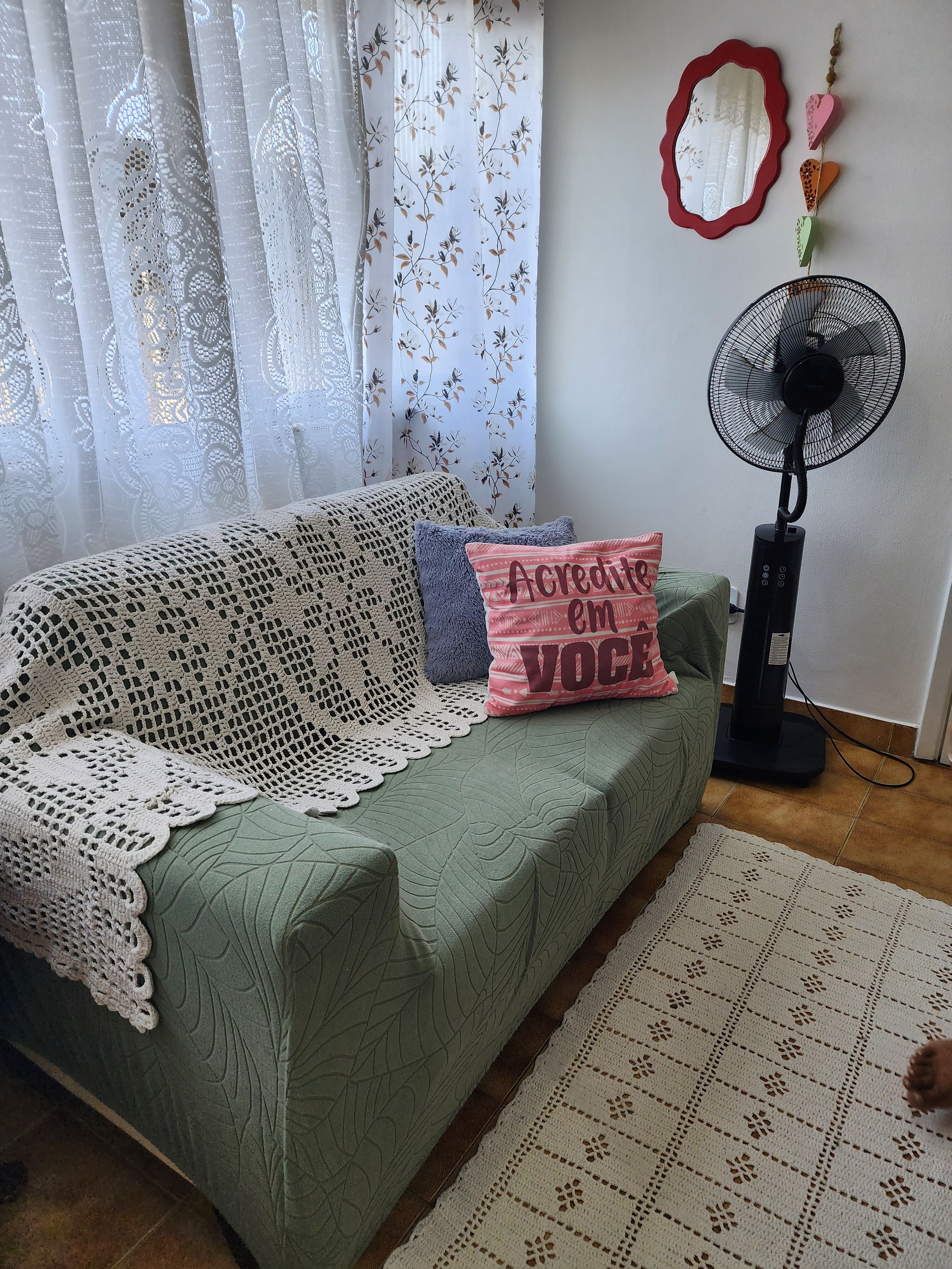 A cozy living room with a green sofa, a white crocheted throw, and a pink pillow with text. A standing fan is on the floor, and a decorative mirror hangs on the wall.