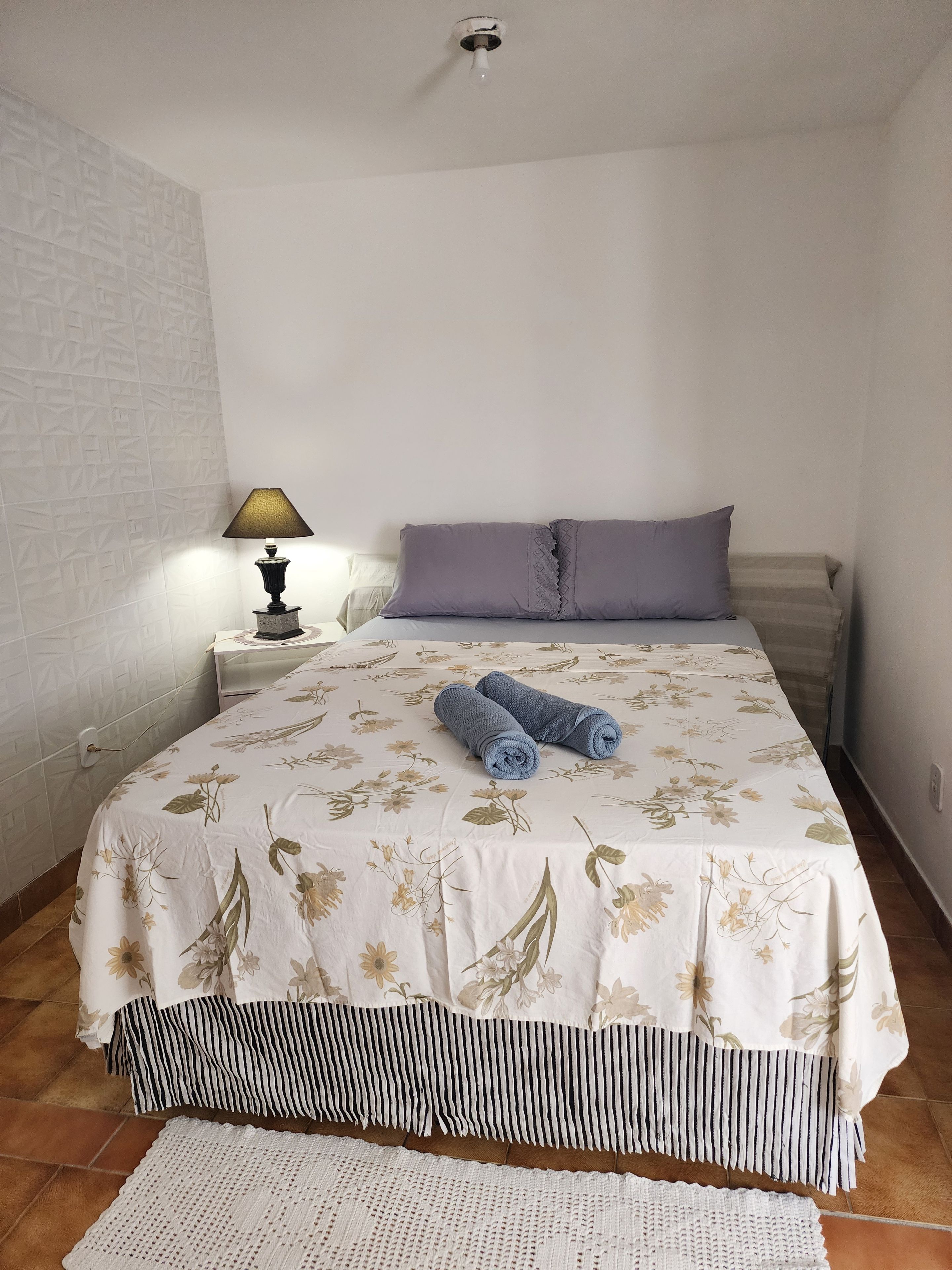 A bedroom with a bed, a nightstand, a lamp, and a rolled towel.