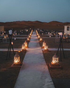 Um caminho no deserto iluminado por lanternas, levando a um edifício com mesas e cadeiras.