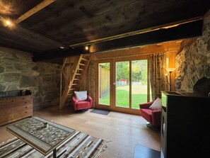Une pièce chaleureuse avec un mur en pierre, des poutres apparentes en bois au plafond, une cheminée, un fauteuil rouge, une commode en bois et une grande fenêtre.