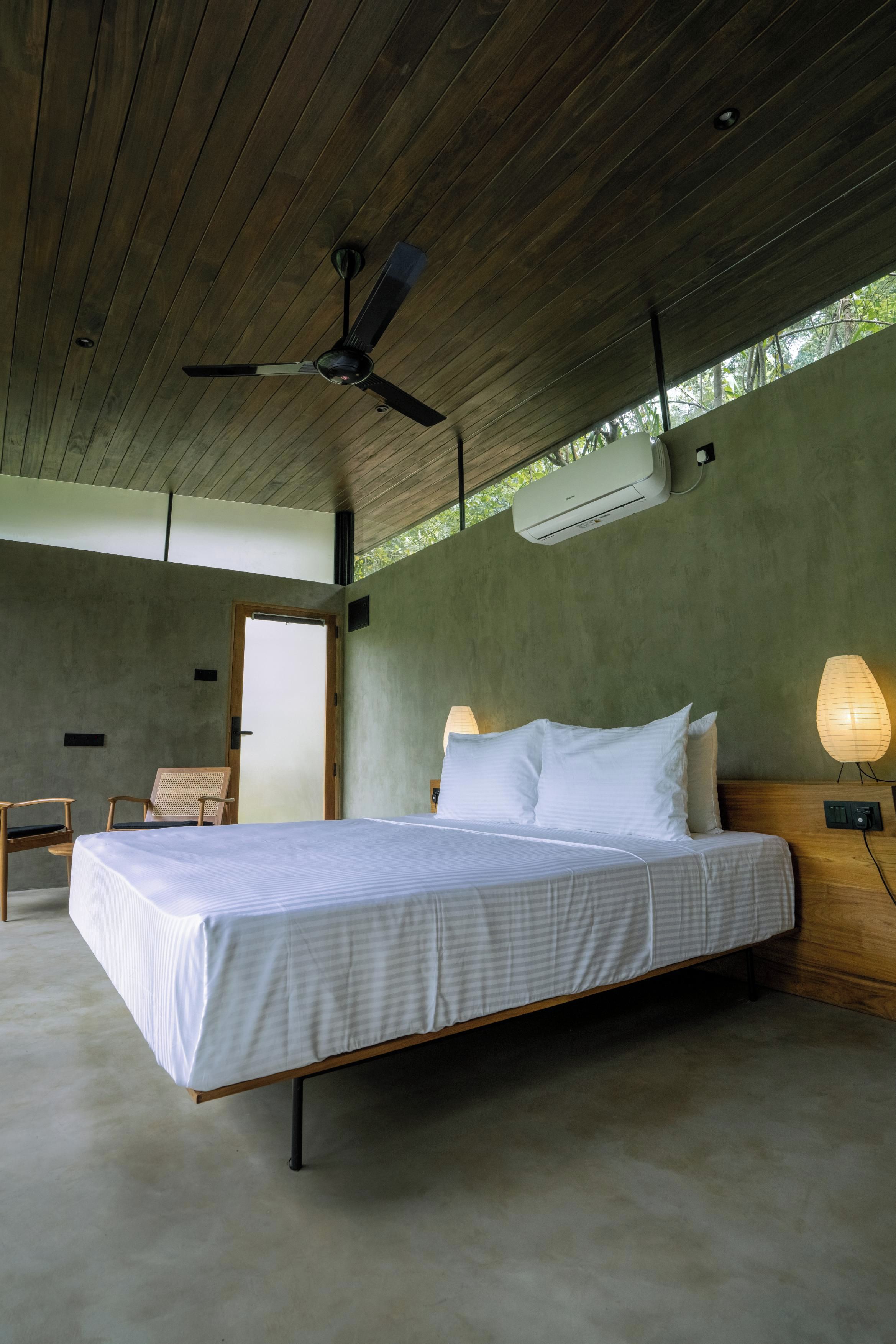 Un dormitorio con cama, ventilador de techo, unidad de aire acondicionado, mesita de noche con lámpara y vistas al exterior.