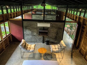 Un espacio de estar al aire libre cubierto con una parrilla, dos sillas con cojines, una mesa redonda de madera y vista a la vegetación a través de la estructura de madera.