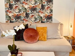 A sofa with a tropical leaf pattern on the wall behind it, a white orchid plant, and a small table with a candle. - Apartment With Garden, Mont-dore (Mont-Dore)