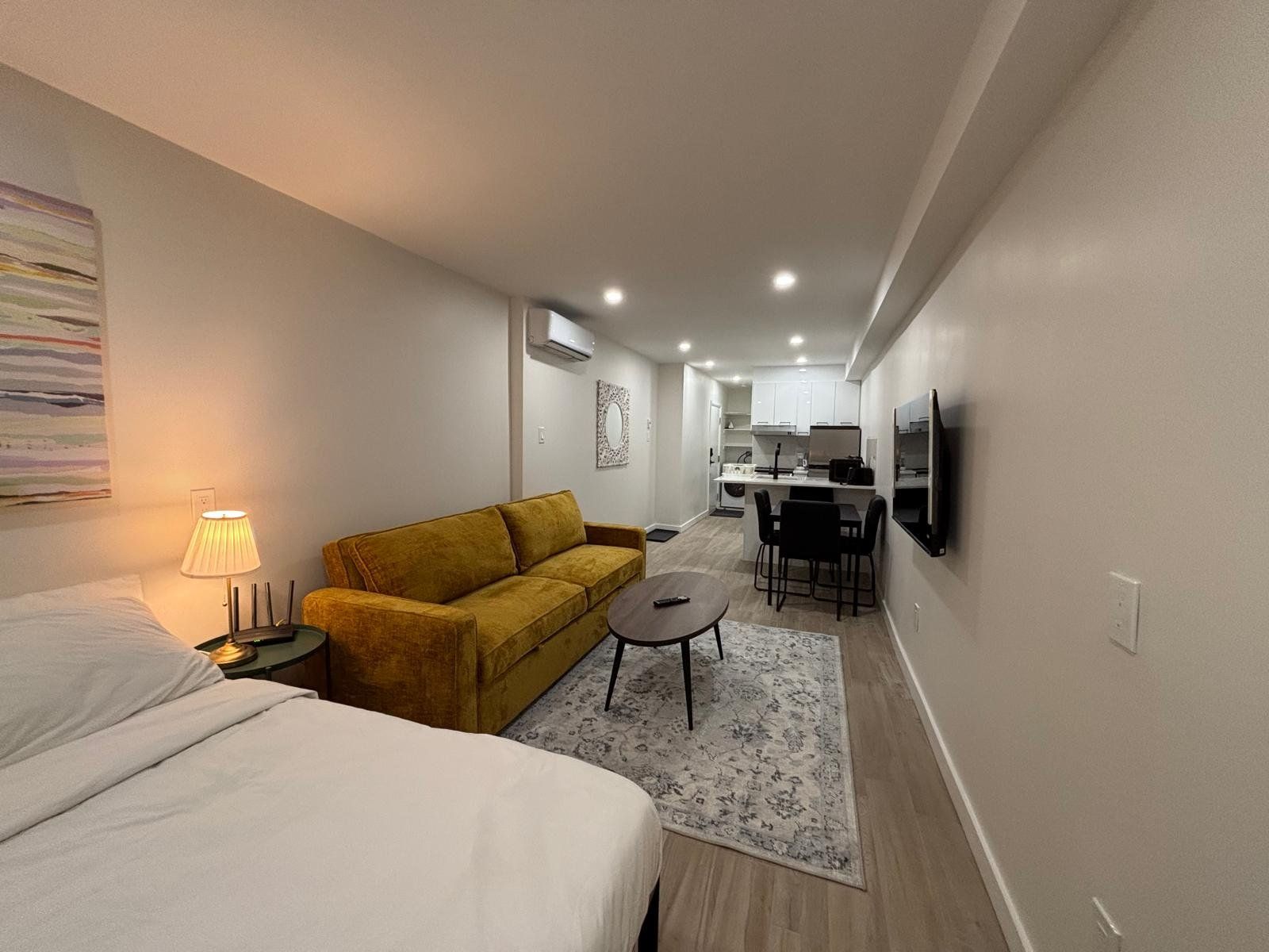 Habitación de hotel compacta con una cama, un sofá, una pequeña mesa y una zona de cocina al fondo.