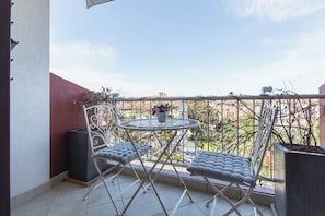 Een balkon met een ronde tafel en twee comfortabele stoelen, een bloempot en een uitzicht op gebouwen op de achtergrond.