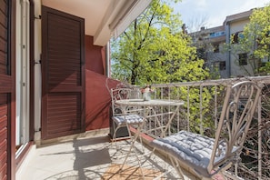 Een balkon met een metalen tafel en stoelen, een gewatteerd bankje en uitzicht op bomen en gebouwen.