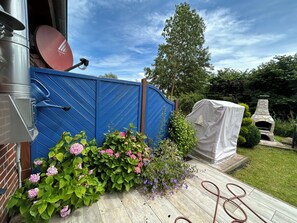 A garden with a blue fence, a stone barbecue grill, and blooming flowers. - Vacation home for up to 5 people—non-refundable, no pets allowed, no free cancellation (Boltenhagen)