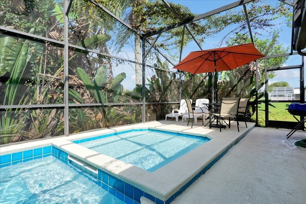 Une piscine extérieure dotée d’une structure en verre, entourée de plantes tropicales et agrémentée d’un parasol rouge.
