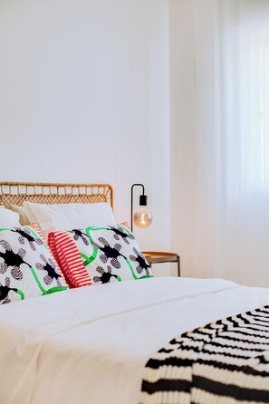 Cama bem arrumada com roupa de cama estampada, mesa de cabeceira com abajur e janela com cortinas leves.