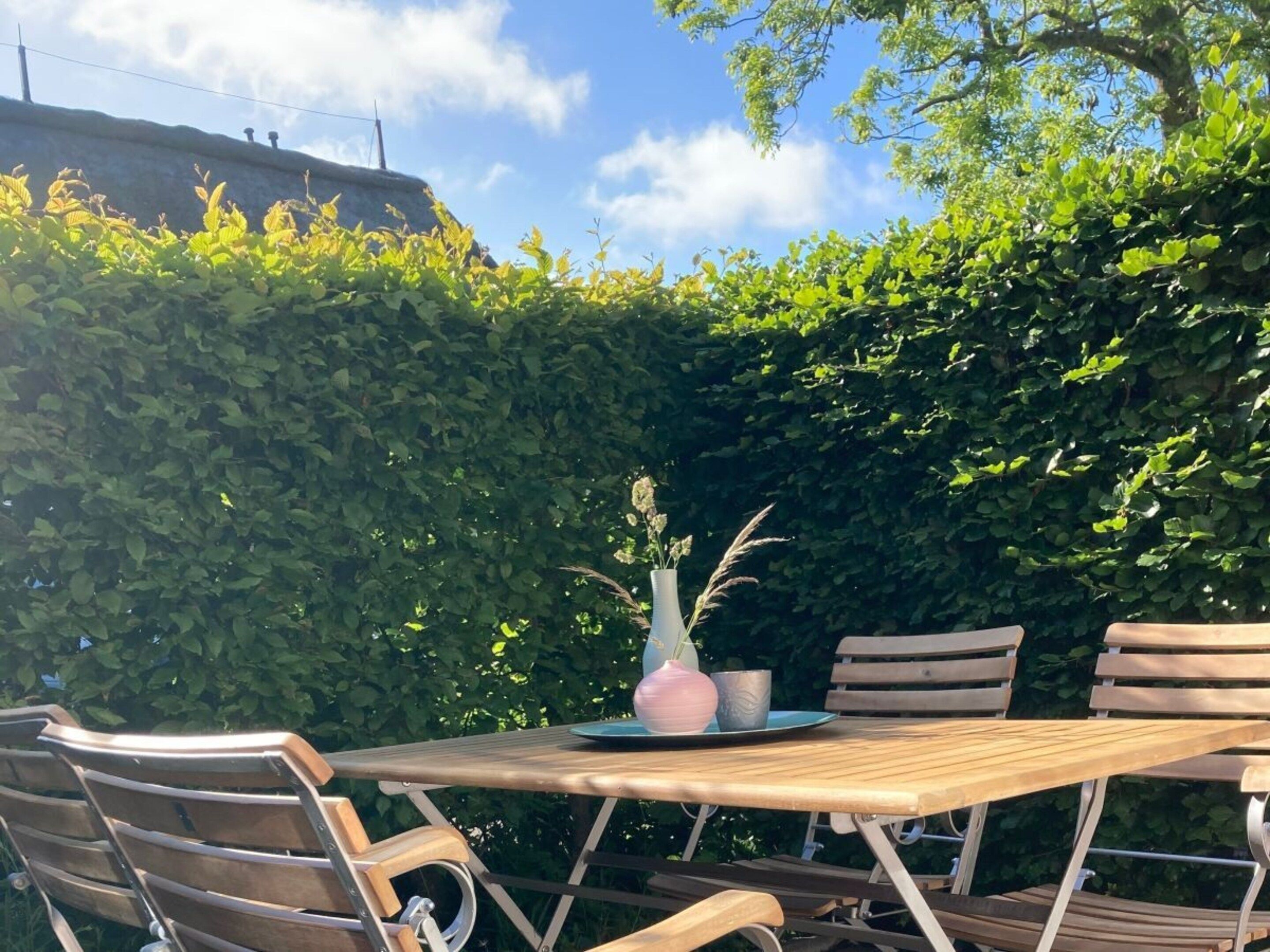 An outdoor wooden table with chairs, a vase with flowers, and a hedge in the background.