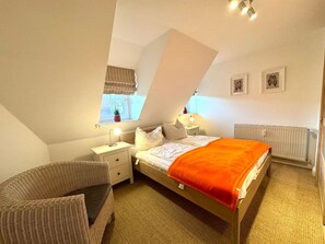 A bedroom with a sloped ceiling, a bed with an orange blanket, a nightstand with a lamp, a chair, and a window with a view. - Holiday Apartment With 1 Bedroom (Borgsum)