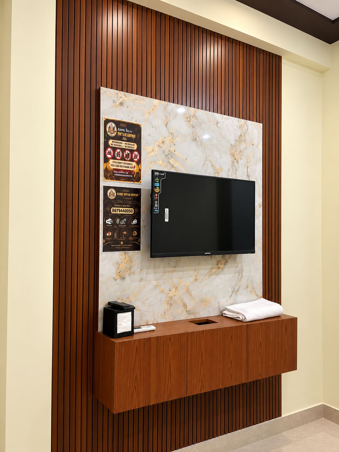 A modern hotel lobby with a marble wall, a flat-screen TV, a wooden reception desk, and informational posters.