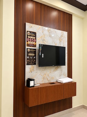 A modern hotel lobby with a marble wall, a flat-screen TV, a wooden reception desk, and informational posters. - HOTEL RADHE SHYAM KRIPAM (Mathura)