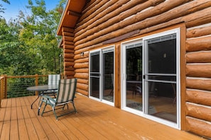 A wooden deck with a table and chairs, large sliding glass doors, and a view of trees. - Handcrafted Log Cabin on 40 Wooded Acres | Near Boyne & Gaylord | Sleeps 6 (Vanderbilt)