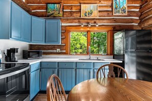 A modern kitchen with blue cabinets, a wooden dining table, and a large window overlooking a forest. - Handcrafted Log Cabin on 40 Wooded Acres | Near Boyne & Gaylord | Sleeps 6 (Vanderbilt)