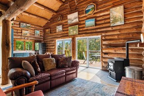 A log cabin interior with a leather sofa, dining table, and a wood-burning stove. - Handcrafted Log Cabin on 40 Wooded Acres | Near Boyne & Gaylord | Sleeps 6 (Vanderbilt)
