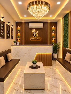 A hotel reception area with a marble reception desk, a chandelier, wall-mounted air conditioning, and a logo for Vaishnavi Grand. - Hotel Vaishnavi Grand 200 m 80 Ghat (Varanasi)
