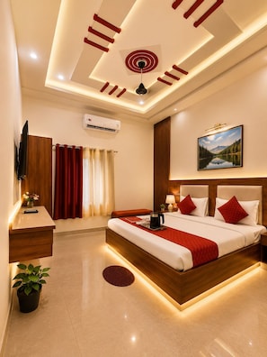 A hotel room with a large bed, bedside tables, a TV, an air conditioning unit, and a painting on the wall. - Hotel Vaishnavi Grand 200 m 80 Ghat (Varanasi)