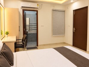 A hotel room with a bed, a desk, a chair, a window with blinds, and a door. - Hotel Vaishnavi Grand 200 m 80 Ghat (Varanasi)