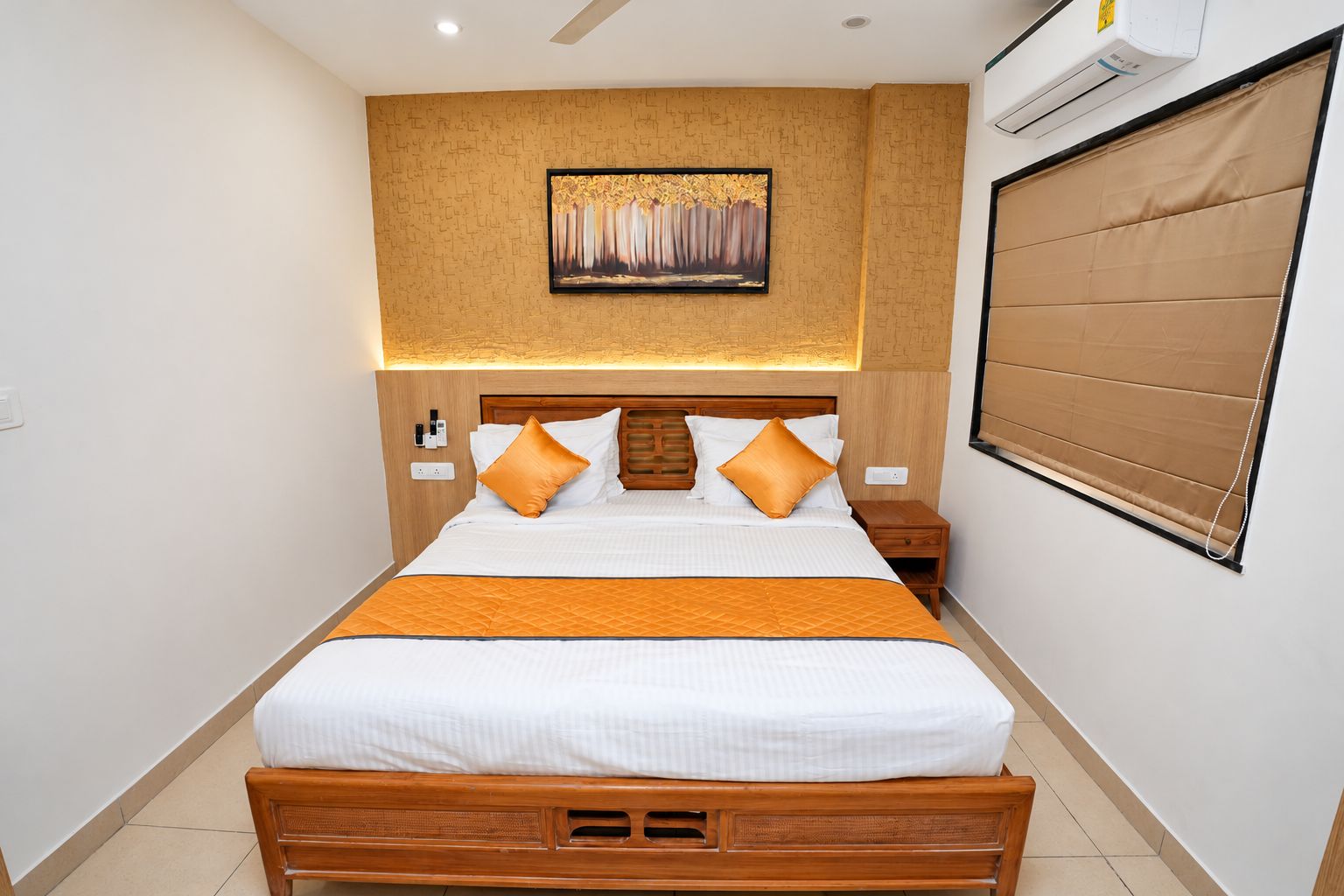 A hotel room with a wooden bed, a headboard with a built-in shelf, a wall-mounted air conditioner, and a large window with a blind.