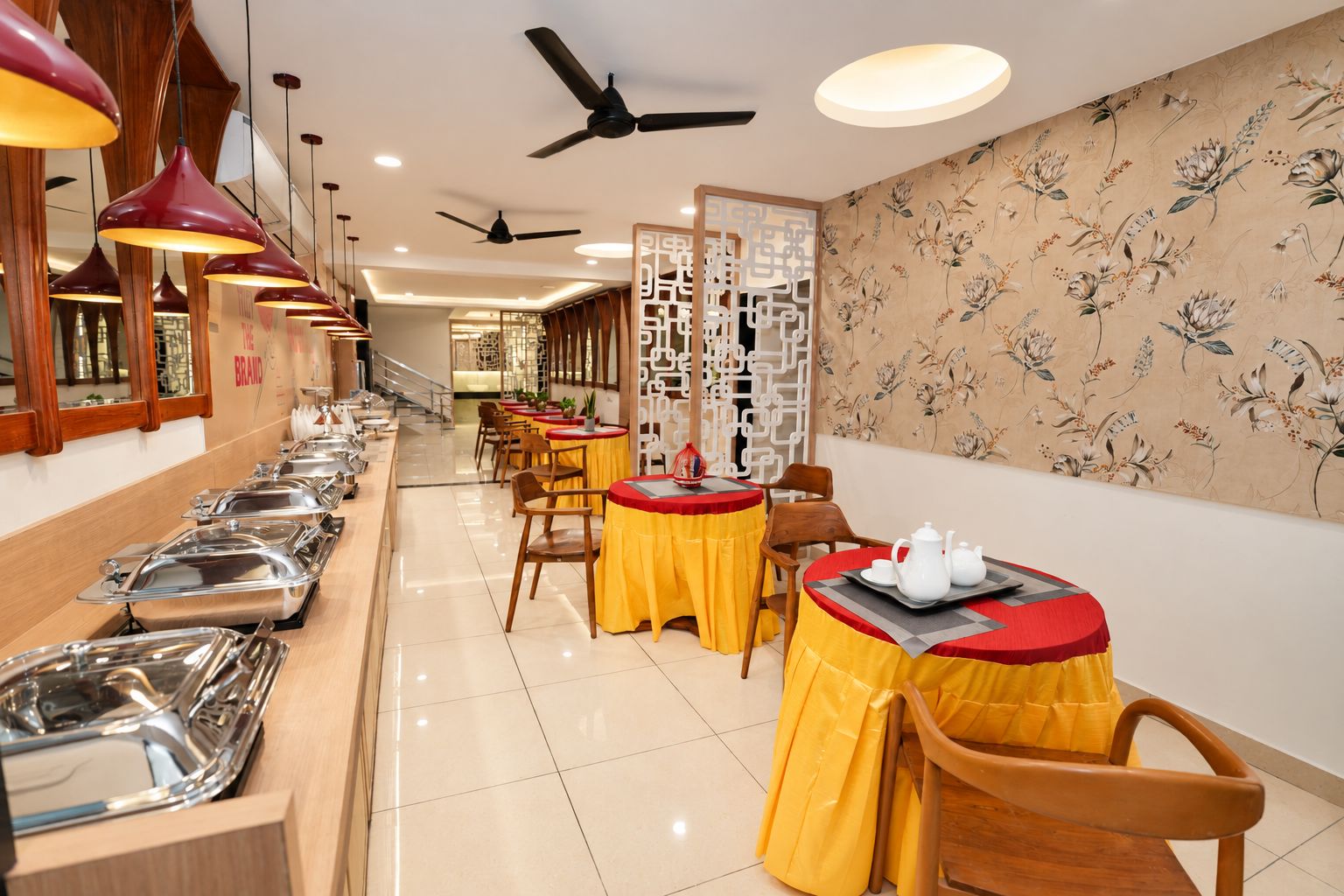 Uma área de buffet com várias estações de servir e mesas, um ventilador de teto e uma parede decorativa com padrões florais.
