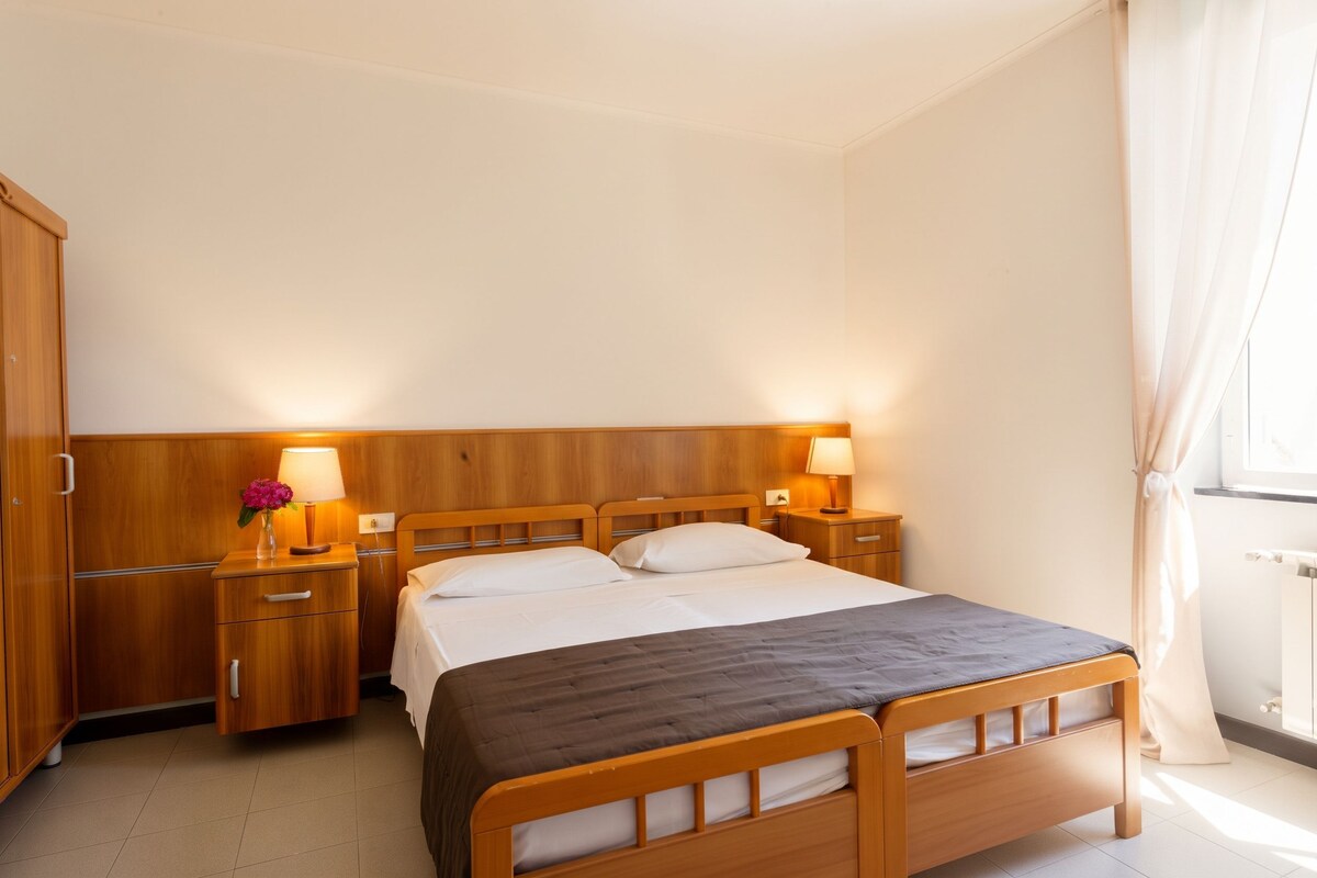 Quarto de hotel com cama de casal, cabeceira de madeira, mesas de cabeceira com luminárias, guarda-roupa e radiador.
