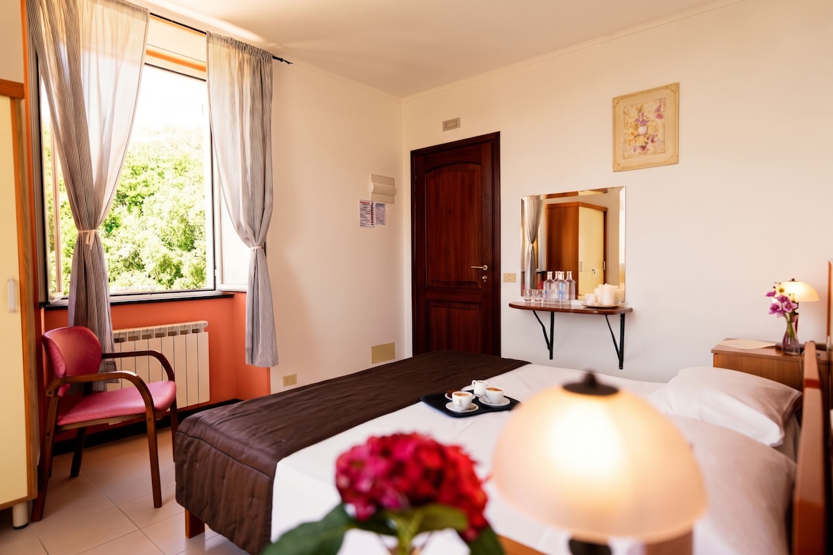 Quarto de hotel com cama, uma cadeira, mesa com abajur e janela com cortinas.
