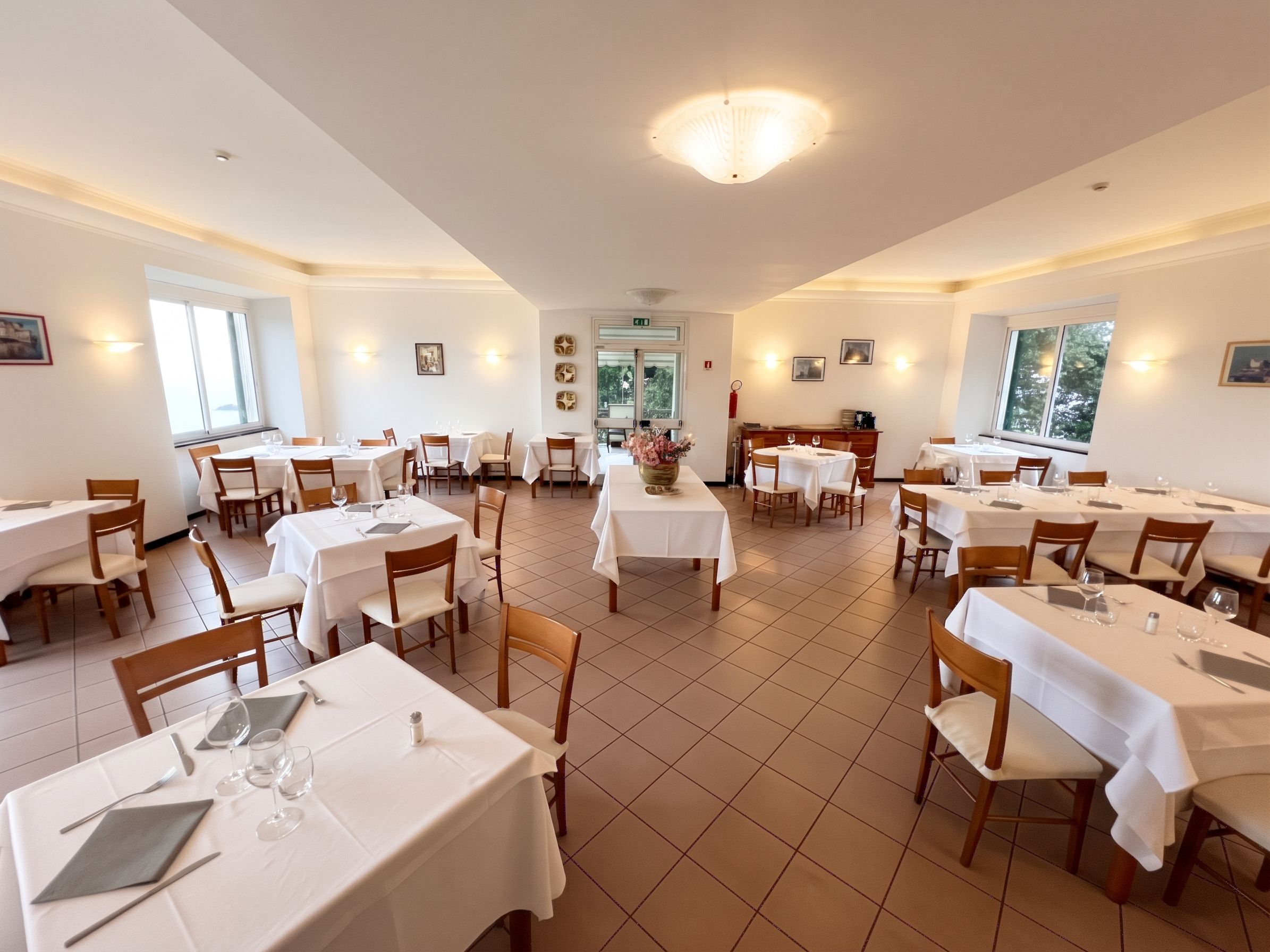 A restaurant with multiple tables and chairs, white tablecloths, and a tiled floor.