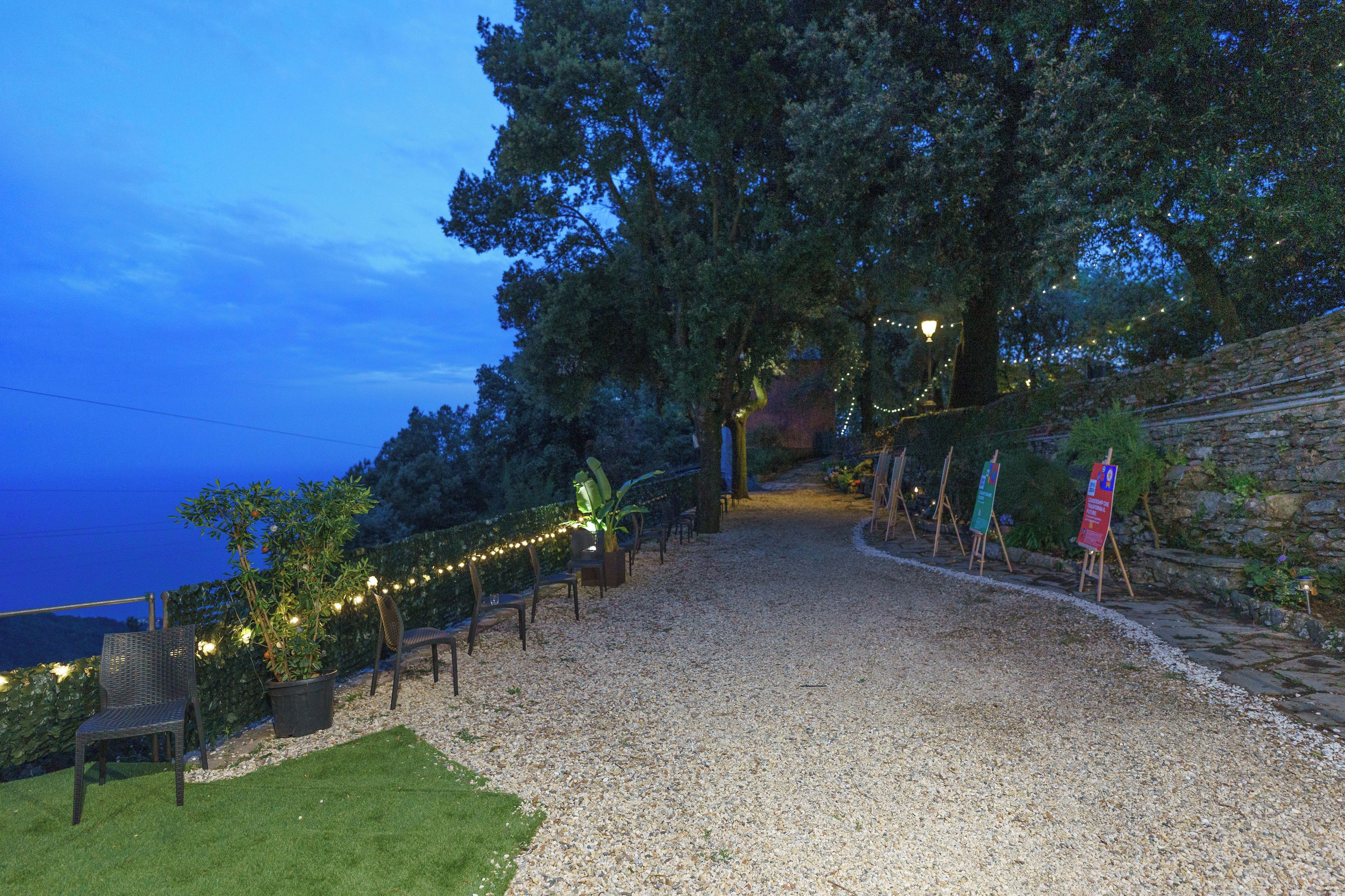 Un sentiero sterrato conduce a una zona con sedute, tavoli, alberi e un muro in pietra.
