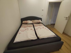 Un llit individual amb un panell de capçal fosc i roba de llit floral a una habitació amb parets blanques i un terra de fusta.