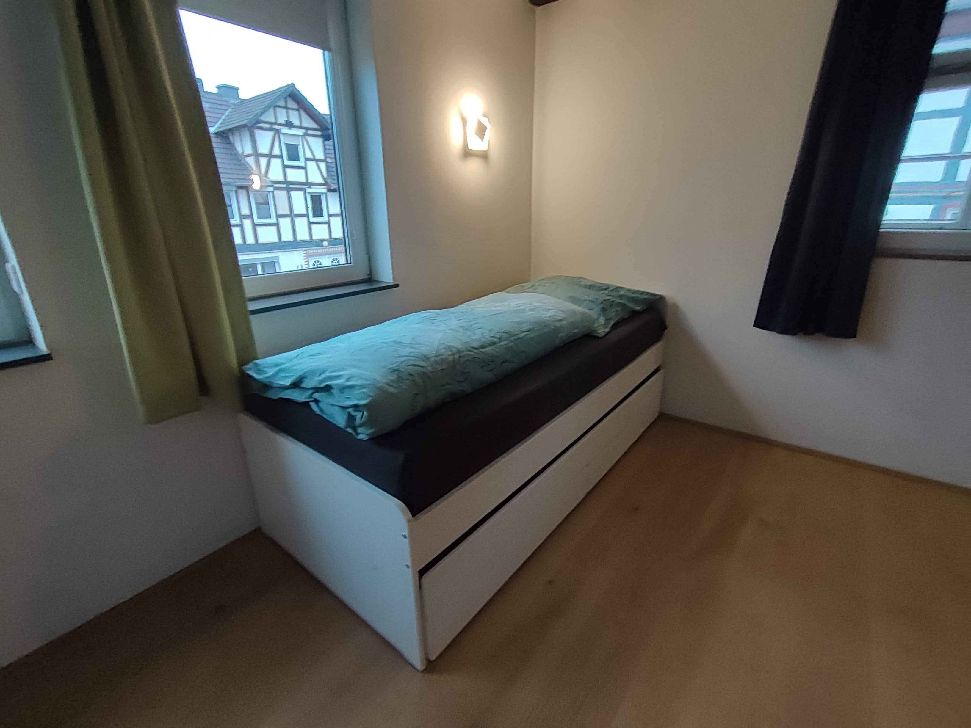 A small, single-bed room with a window, a wall-mounted light, and a curtain.
