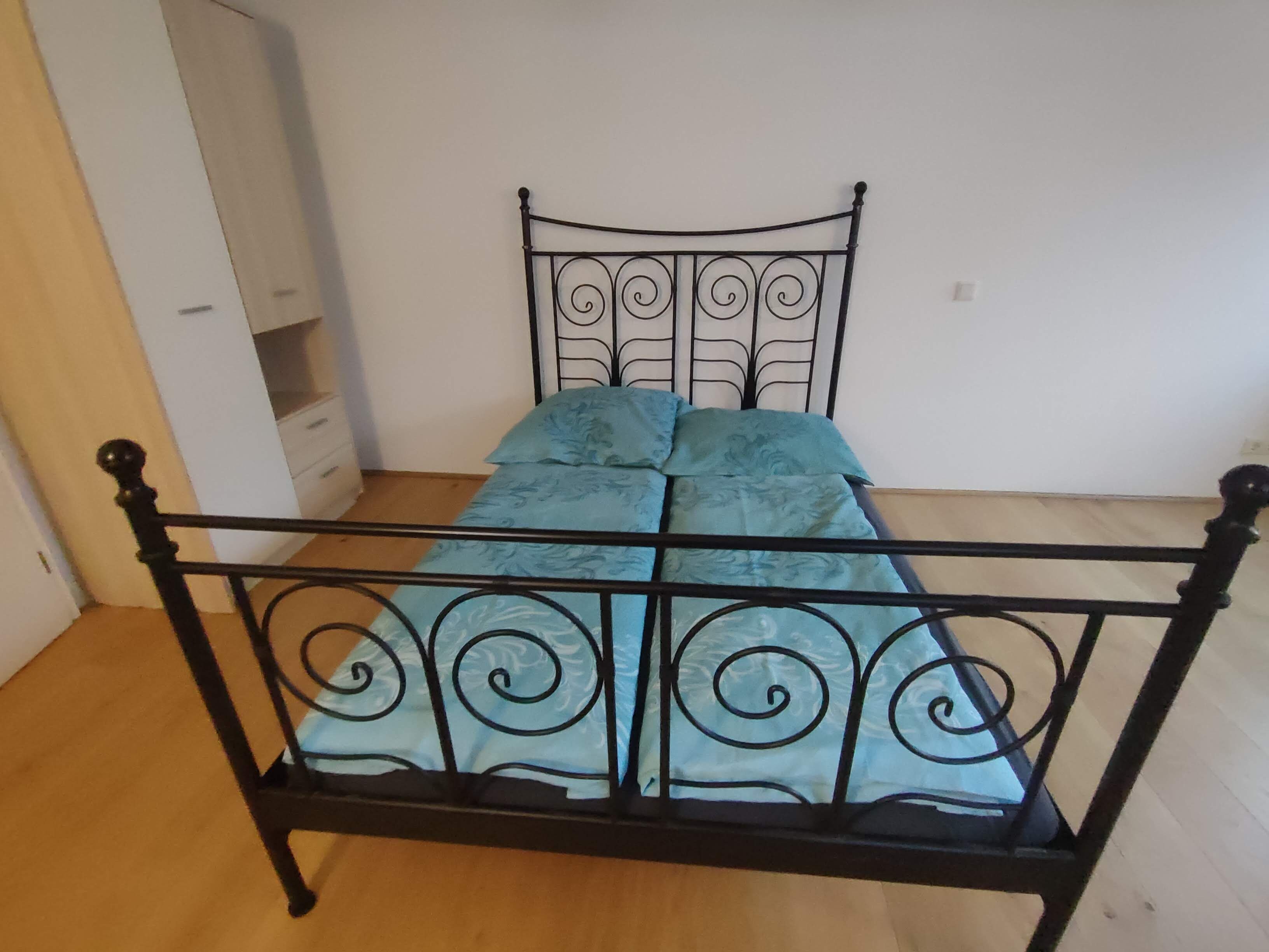 Ein Bett mit Metallgestell und einem gemusterten Bettbezug in einem Zimmer mit Holzboden und weißen Wänden.