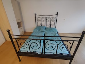 Una cama con estructura metálica y colcha estampada en una habitación con piso de madera y paredes blancas.