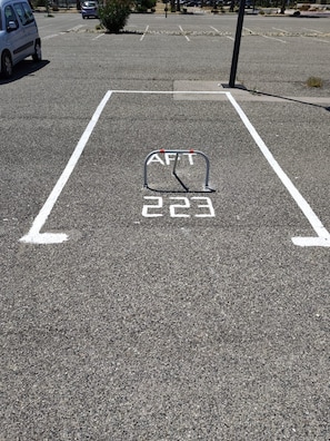 Ein Parkplatz mit weißer Markierung für einen Stellplatz, einer Metallsperre mit dem Aufdruck 'AFT 223' und einem kleinen Baum im Hintergrund.