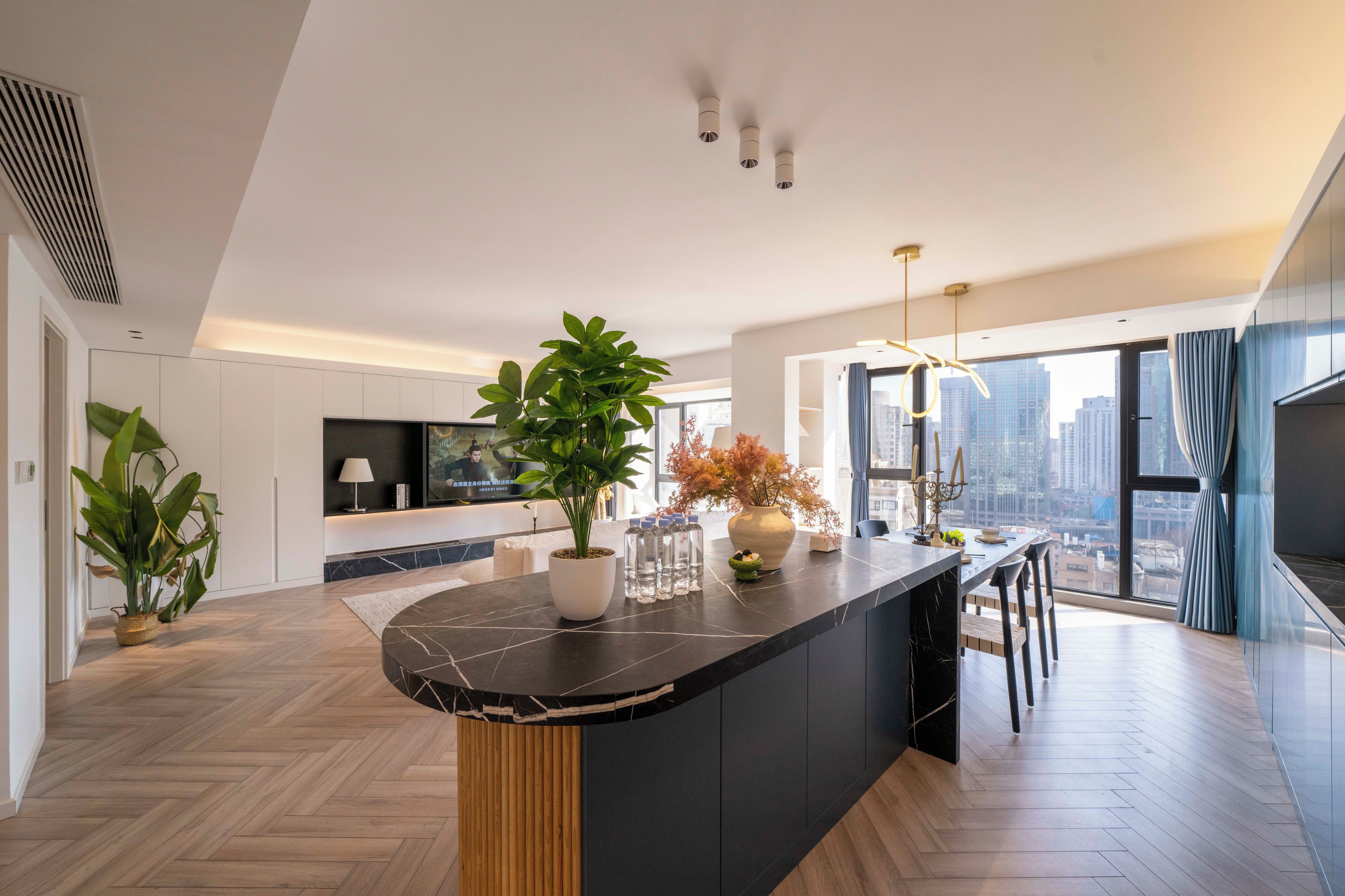 A modern kitchen with a marble countertop, a large potted plant, and a city view through large windows.
