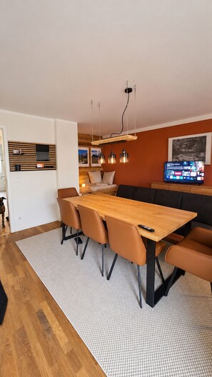 Een moderne woonruimte met een houten eettafel, lederen stoelen, een bank en een televisie waarop een spel wordt getoond.