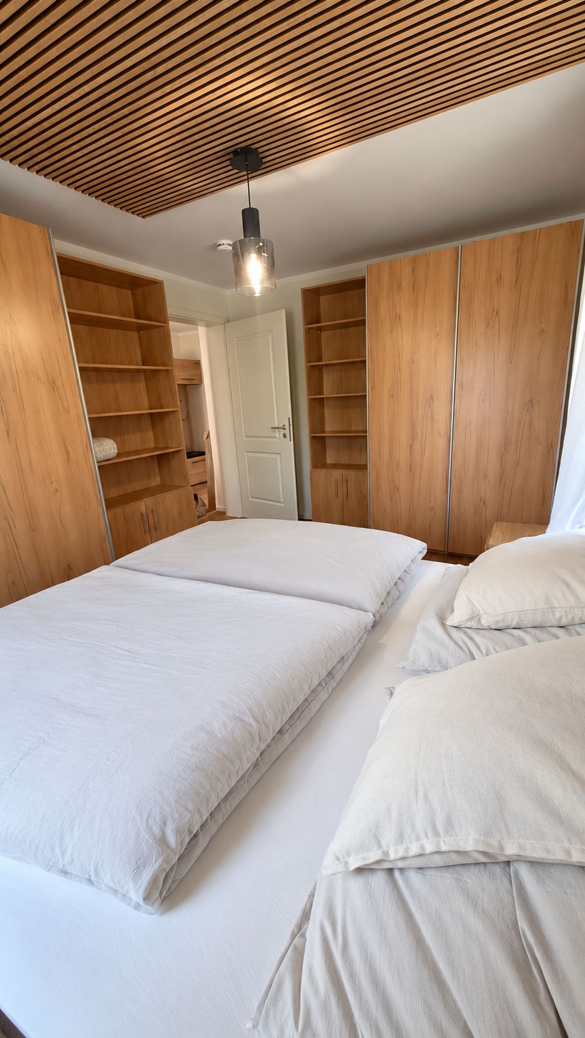 A bedroom with a wooden slatted ceiling, a bed with white bedding, and built-in wooden shelving units.