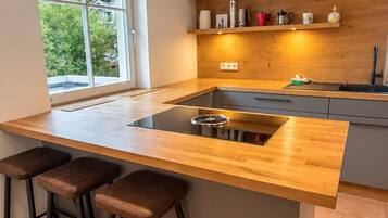 A modern kitchen with a central island, bar stools, pendant lighting, and a window with a view of greenery.