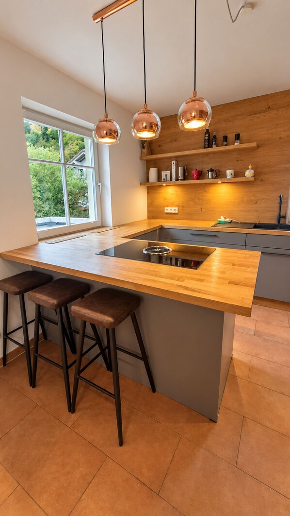 Een moderne keuken met een centraal eiland, barkrukken, hanglampen en een raam met uitzicht op groen.