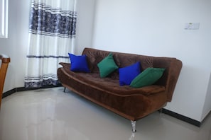 Um sofá marrom com quatro almofadas em um quarto com parede branca e janela com cortinas estampadas.