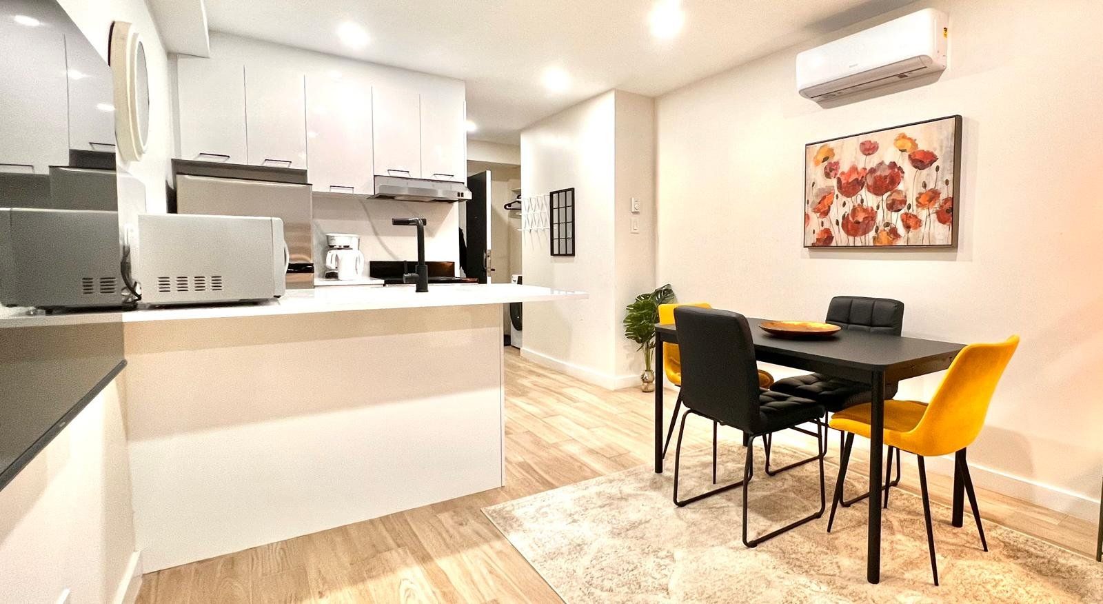 Una cocina moderna con armarios blancos, una isla central y un comedor con mesa negra y sillas amarillas.
