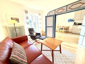 A living room with a sofa, a chair, a coffee table, a radiator, a window with curtains, and a kitchen area visible through an open door. - Garden Apartment in the Muschel House (Nieblum)