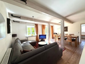 A living room with a grey sofa, a flat-screen TV, a dining area with wooden furniture, and a window with curtains. - Holiday Home in Oldsum With Swimming Pool (Oldsum)