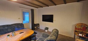 Ein Wohnzimmer mit einer Couch, einem Tisch mit Fernbedienung, einem Fernseher und einem kleinen Regal mit Gegenständen.