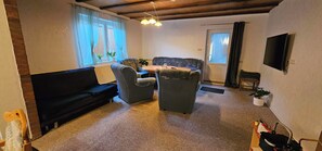 A cozy living room with a dark sofa, two armchairs, a small table, and a television mounted on the wall. - Vacation Home "Im alten Kiosk" - Relaxation Right on Lake Idasee (Ostrhauderfehn)