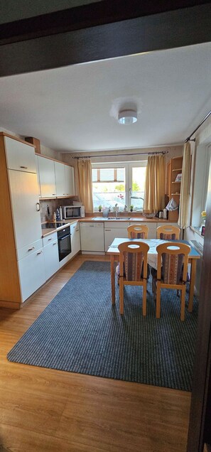 A compact kitchen with white cabinets, a microwave, and a sink. There is a dining area with a table and chairs. - Vacation Home "Im alten Kiosk" - Relaxation Right on Lake Idasee (Ostrhauderfehn)