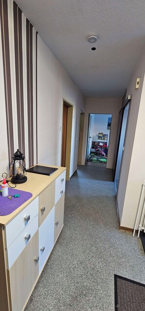 Ein Flur mit Schreibtisch, Schubladen, Heizkörper und Blick in einen Raum mit Bett und Fenster.