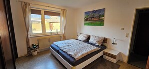 Ein Schlafzimmer mit einem Bett, einem Fenster mit Vorhängen, einem Heizkörper, einem kleinen Nachttisch und einem Bild an der Wand.