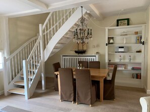 A dining area with a wooden table and chairs, a chandelier, a staircase, and a bookshelf with books and decorative items. - 5 in the Deichhof House (Dunsum)