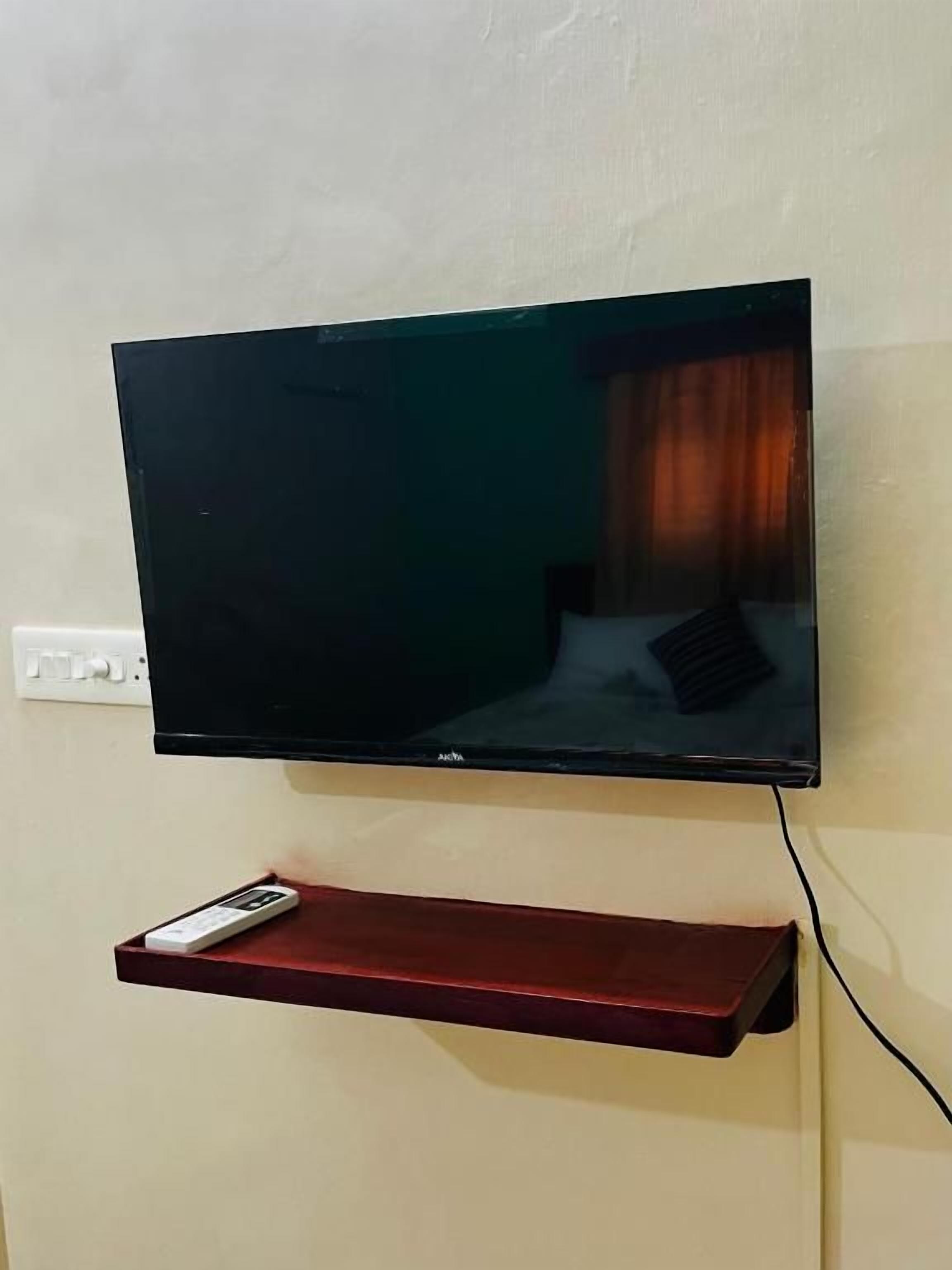 A flat-screen TV mounted on a wall with a remote control on a red shelf below.